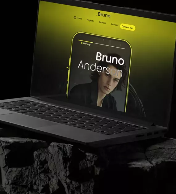 Laptop on textured rock surface displaying Bruno Anderson portfolio website with yellow-green background and iPhone mockup showing contact section