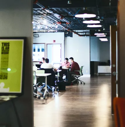 View through a doorway into a dimly lit open-plan office with people working at desks under pendant lights