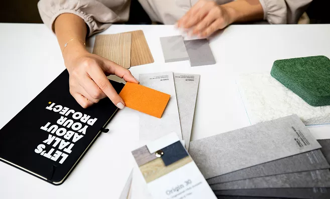 Designer's hands arranging material swatches on a white table beside a black notebook reading "Let's Talk About Your Project."