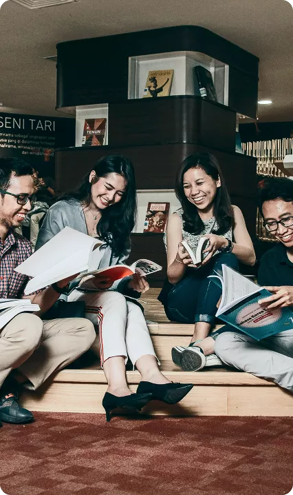 Four young adults sitting on wooden steps in a library, laughing and reading colorful books together