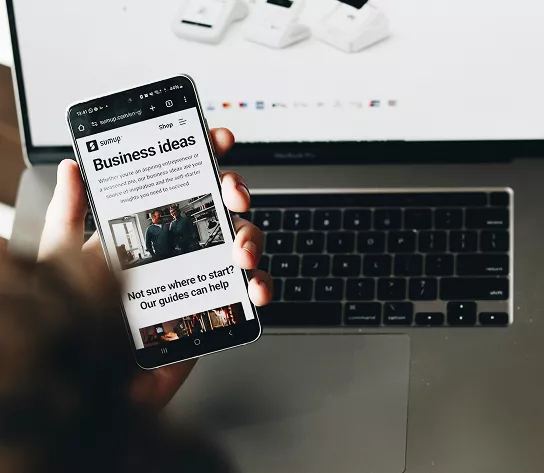 A hand holds a smartphone displaying a SumUp 'Business Ideas' webpage, with a MacBook laptop and white card readers visible in the background.